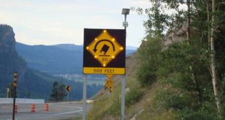 Figure 17. Photo. Dynamic warning sign showing activated truck rollover warning system. This photo shows a square black sign with a yellow strip at the bottom with the legend '500 Feet.' The large, top, black portion has a yellow diamond-shaped sign with flashing lights along its edge. The diamond-shaped sign has black symbols showing the rear view of a tilted large truck; above it is a semicircular arrow at the top pointing left, and below it is '20.' The sign conveys a risk of rollover for trucks.