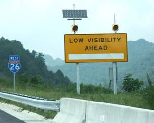 Figure 22. Photo. Weather advisory system using a static sign with beacons. This photo shows a sign just off the roadway of I-26 West. The orange, rectangular sign has two flashing beacons and says 'Low Visibility Ahead.'