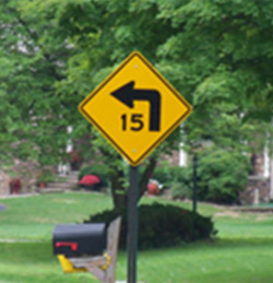 Photo. A yellow advisory sign with a black speed advisory of 15 MPH and curve warning. 