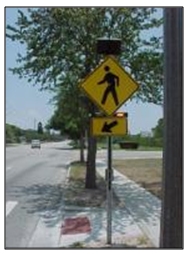 Photo ofa crosswalk warning sign at the right of a roadway preceding a crosswalk. The warning sign features a pair of rectangular rapid-flashing beacons below the sign but above an arrow placard indicating the location of the crosswalk.