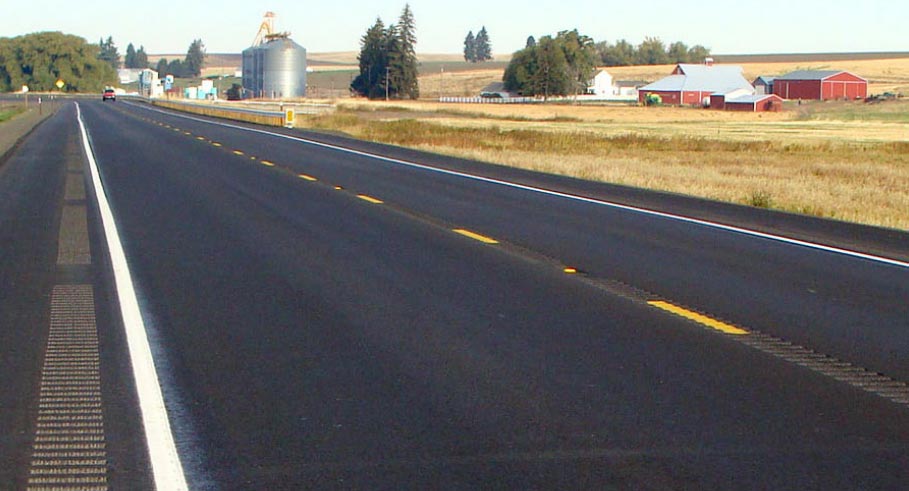 Photo: This photograph, taken along the edge line of a two-lane undivided roadway, shows longitudinal rumble strips present in the center and along the paved shoulder of the roadway. The roadway is located in a level, rural setting, and the roadway is marked with a dashed yellow centerline and solid white edge lines.