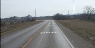 Photo.  A curve approach on a two lane roadway is shown with pavement markings that say CURVE-50-MPH.