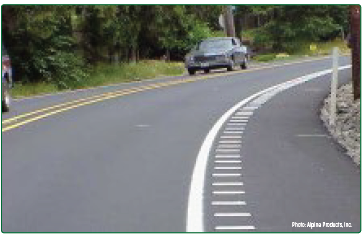 A curving roadway with raised white strips on the shoulder just outside the travel lane that function as rumble strips.