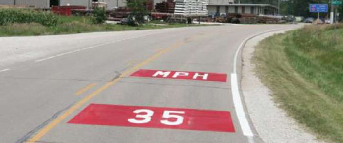 Figure 5-7 Horizontal signing at community entrance in Dexter, IA [Treatment authorized with MUTCD experimental waiver] (Image Source: Neal Hawkins). This photo shows an experimental treatment with 35 MPH painted in red on the road. Note: The red background coloring is not MUTCD compliant, and should only be installed after receiving approval from FHWA through an experimentation waiver.