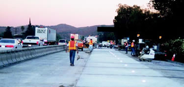 workers wearing orange vests and hardhats beside a concrete traffic barrier