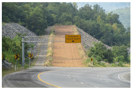 Figure 8. Photo. Truck escape ramp. This photo shows a truck escape ramp with a sign that says 'Runaway Truck Ramp.' The roadway curves to the right, while the ramp continues straight and dissipates trucks' energy through gravitational deceleration (a sharp incline) and rolling resistance (a gravel bed).