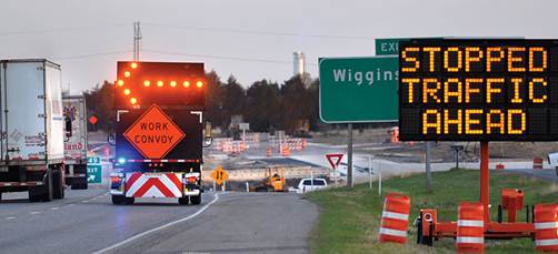 Figure 21. Photo. Dynamic queue detection warning sign in a work zone. This photo shows a work zone and warning signs. A work truck is stopped in the rightmost travel lane and bears an orange diamond-shaped sign saying 'Work Convoy' and above it a flashing orange arrow pointing left. A dynamic message sign is just off the roadway to the right, along with orange safety cones, and says 'Stopped Traffic Ahead.'