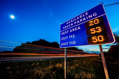 Figure 24. Photo. SmartPark in Michigan. This photo shows a roadway at dusk with blurred traffic and a rectangular blue sign just off the roadway to the right. The sign says 'Available Truck Parking' at the top. Below it are three options with a dynamic message listing the number of available spaces: 'Rest Area 20,' 'Exit 112 53,' and 'Exit 110 50.'