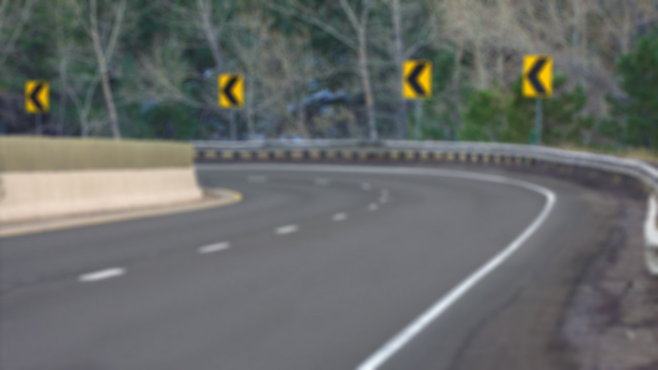 Blurry photo of horizontal curve on a rural road with guard rail and four yellow chevron signs delineating the curve.