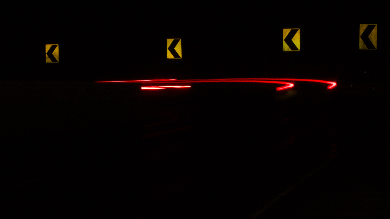 Night time image of a horizontal curve. The image is black except for four chevron signs reflecting the headlights of a car going through the curve. There are also red streaks showing the tail lights of a car going through the curve.