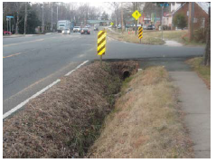 A roadway drops off into a drainage ditch that runs under a cross-street that serves as the entrance to a residential neighborhood.
