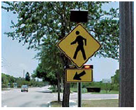 A rectangular flashing beacon above a pedestrian crossing sign.