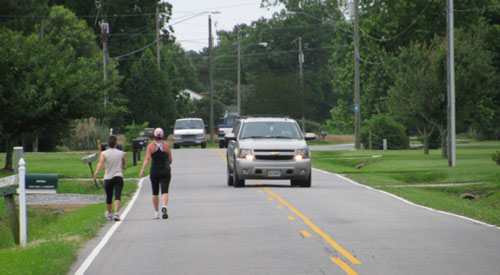 Photo.  A rural two-lane  road with no shoulder .  Two pedestrians walk on the far left, facing traffic.  An on-coming vehicle has to encroach on the opposite travel lane to avoid the pedestrians.