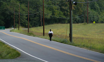 Photo. A pedestrian walking on the right side of the road, the same direction as motorized traffic, on a road with no shoulder.