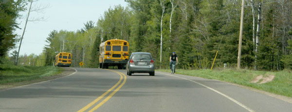 Photo.  A two lane rural road with a shoulder only on the right side.  A cyclist is traveling against traffic on the shoulder.  