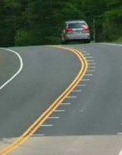 Photo. A rural two-lane road with optical speed bars on the right lane as it approaches a horizontal and vertical curve. The bars are short white lines on either side of the lane and are placed closer together approaching the curve to give the driver the illusion that they are driving faster. 