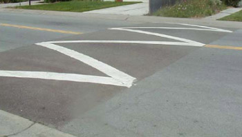 Photo. A speed hump, which is a slightly raised section of roadway that is a couple of feet wide and has a zig zag white pavement marking on top. 