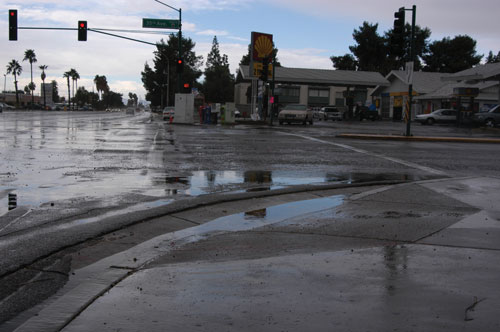 Photo. This photo shows a street corner where water is standing on the roadway and along the curb.