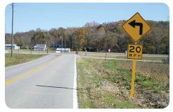 Photo of a curve warning sign in advance of a 90 degree curve. The sign also indicates the maximum safe speed limit on the curve is 20 mph.