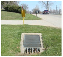 A drainage pipe that bisects a driveway and has a concrete sleeve and grating covering the opening to prevent vehicles from dropping into it.