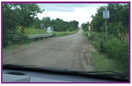An unpaved rural roadway running over a narrow, single-lane bridge with guard rails on either side.