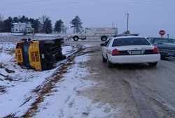 This is a photo of a traffic crash along a rural roadway. A school bus has run off the road and overturned on its side in snowy conditions. A police car and ambulance also are shown.