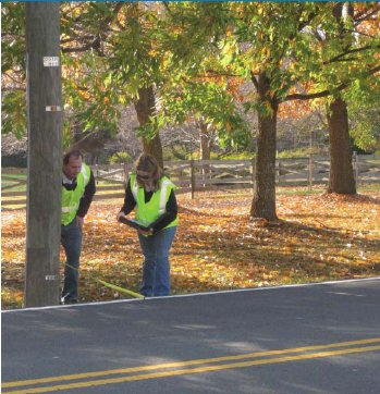 Photo of a man and woman in retroreflective clothing measuring the distance from a utility pole to the edge of a raised roadway in a rural area and recording the data on a clipboard.