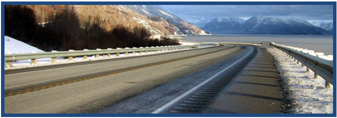Photo features a mountainous area separated from a wide body of water by a curving roadway with snow on the unpaved areas. The road features centerline and edgeline rumble strips and stripes.
