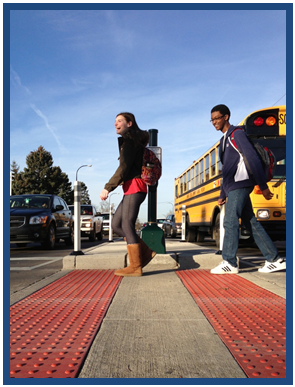 Photo of school children walking in front of a bus in a crosswalk cut through that features strips of truncated domes placed in line with the edges of the median cut-through area to indicate to sight impaired users that they are entering the roadway.