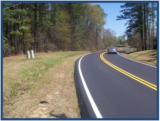 Photo of freshly laid asphalt on a two-lane roadway featuring the safety edge. The roadway curves through a wooded area, but has wide, grassy shoulders.