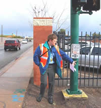 A man in an orange safety vest presses the call button at a pedestrian crossing.
