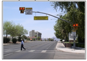 Photo of a pedestrian walking in a crosswalk at an intersection that features the HAWK system, with a pole-mounted set of dual solid red lights and a walking person symbol to indicate the presence of a pedestrian and that drivers should stop to allow the pedestrian to cross.