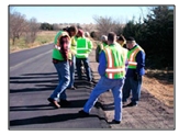 Photo of a group of men wearing Class 2 retroreflective vests standing on a newly paved but unmarked roadway examining the edgeline.