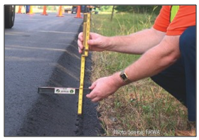 Photo of a roadway worker holding a horizontal level and a verticle measuring stick at the edge of a newly laid roadway, depicting the 30 degree angle that results from application of the Safety Edge.