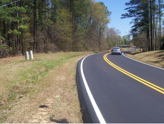 Freshly laid asphalt on a two-lane roadway featuring the safety edge. The roadway curves through a wooded area, but has wide, grassy shoulders.
