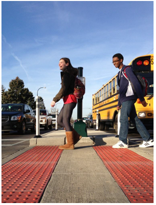 School children walking in front of a bus in a crosswalk cut through that features strips of truncated domes placed in line with the edges of the median cut-through area to indicate to sight impaired users that they are entering the roadway.