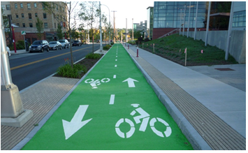 A two-lane roadway with double yellow center lines that is bordered on the right by a wide but street-level brick median featuring pole-mounted luminaires and plantings. The median area serves to separate the roadway from a set of bike lanes, one in each direction, painted a bright green color with dashed white lines between the two lanes, which feature directional arrows and bicycle icons. The bike lanes are bordered on the right by the raised curb of a pedestrian sidewalk.