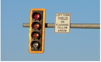 A signal head mounted on a mast arm over a roadway. The signal head features a retroreflective backplate that surrounsds the signal head, improving its visibility at night. A sign next to the signal head reads 'Left turn yield on flashing yellow arrow.'