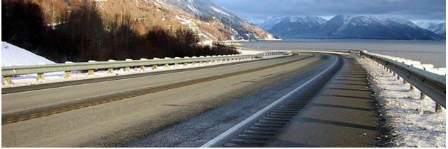 A mountainous area separated from a wide body of water by a curving roadway with snow on the unpaved areas. The road features centerline and edgeline rumble strips and stripes.