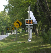 A speed warning device is posted in advance of a rural curve to warn drivers approaching at a dangerous speed to slow down.