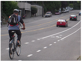 A cyclist uses a dedicated bike lane that is part of a road diet reconfiguration that includes two through lanes, a two-way left turn lane, parallel parking on both sides of the roadway, and the dedicated bike lane.