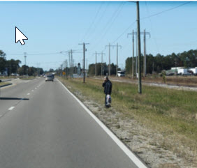 A pedestrian stands on the unpaved roadside of a two-lane roadway waiting to make a mid-block crossing.