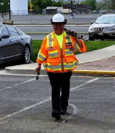 This photo shows a woman wearing retroreflective gear carrying a tripod over her shoulder as she walks across a parking lot.