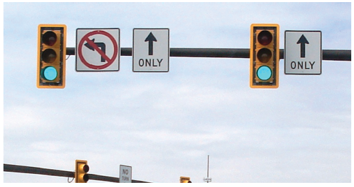 Figure 24. A picture of two traffic signal heads mounted on a mast arm.  Both signal heads have a yellow retroreflective strip around their outer borders.  To the right of both signal heads is a through-only Mandatory Movement Lane Control (R3-5a) sign; a symbolic no-left-turn (R3-2) sign is next to the signal head on the left.
