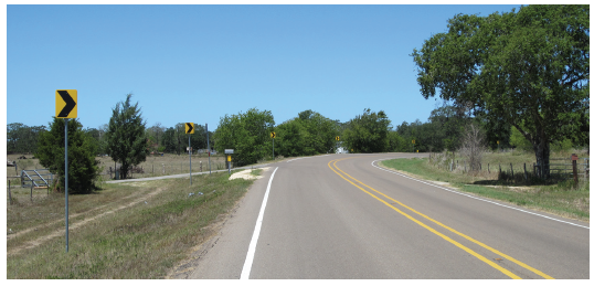 Figure 51. A picture of a horizontal curve to the right on a two-lane rural highway, with post-mounted chevrons on the left roadside, painted white edgelines, and raised pavement markers in the centerline.