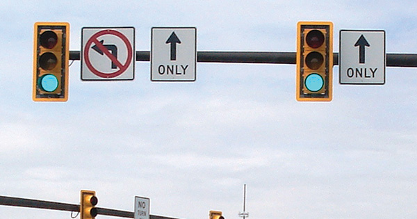 Figure 24. A picture of two traffic signal heads mounted on a mast arm.  Both signal heads have a yellow retroreflective strip around their outer borders.  To the right of both signal heads is a through-only Mandatory Movement Lane Control (R3-5a) sign; a symbolic no-left-turn (R3-2) sign is next to the signal head on the left.