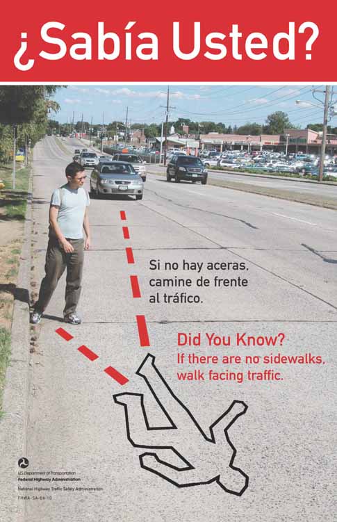The photograph shows a wide suburban road with cars and a male pedestrian on the side of road with his back to traffic. There is no sidewalk. Vehicles approach him from behind. Dotted lines that arrive at a body outline indicate where the paths of a car and the pedestrian would meet if they continue on their present paths. The English text reads: Did you know? If there are  no sidewalks, walk facing traffic. The Spanish tect reads: ¿Sabía Usted? Si no hay aceras, camine de frente al tráfico. 