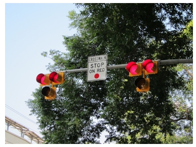 Photo of two High intensity Activated crossWalK (or HAWK) beacon heads suspended on a mast arm. In this photo, each beacon head has two lit red lenses above a single unlit yellow lens. A sign between the two beacon heads warns drivers of a crosswalk and that they should stop on red.
