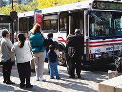 This photo shows people standing in line to board a bus at a designated bus stop. The bus stop has a sign and a shelter.