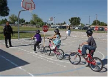 Photo of three children on bikes on a basketball court with a police officer directing them through a mock intersection.